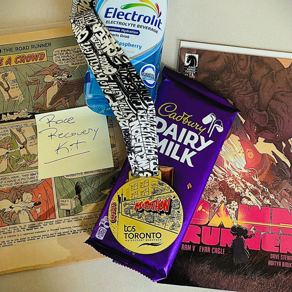 A post-race recovery kit on a desk including the marathon medal, chocolate bar, hydration drink, and a note labeled ‘Race Recovery Kit’ beside a vintage comic strip.